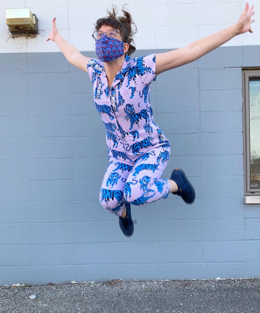 A white woman with brown hair wearing a pink jumpsuit with blue tigers is caught mid-jump with her arms flung wide.